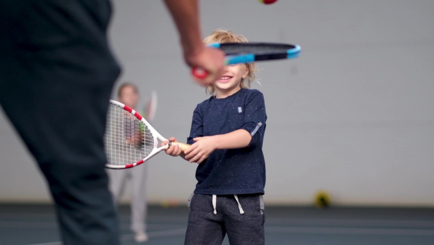 A man coach teaches a little boy to play on an indoor court. A professional tennis instructor throws the ball to hit the racket to the kid, how to return the ball with the racket Royalty-Free Stock Footage #1059976229