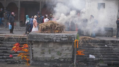Pashupatinath, Kathmandu, Nepal. 12-15-2019. General view of body burning at Pashupatinath temple ghats while family watch in the backgroung and people pass by.