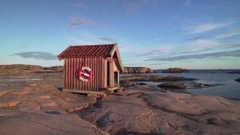 Tiny cabin at coast in Sweden. Fishing hut with life ring safety buoy on red wooden facade. Swedish Bohuslän coastline on evening. cottage on granite rock island. fisher nets 