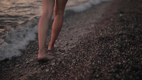 Slim female legs and feet walking along sea water waves on beach. Pretty woman walks at seaside surf. Splashes of water and foam in slow motion. Girl after bathing in ocean go on shore.