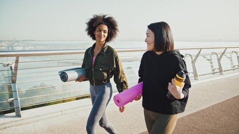 Sport and friendship. Happy asian and african american women going to yoga class, talking on walk, holding fitness mats, slow motion