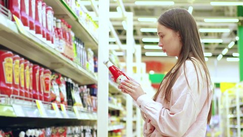 Young woman, customer, is selecting a ketchup sauce in grocery department of hypermarket. She comparing trademarks, prices, reading labels and ingredients. Close up, slow motion