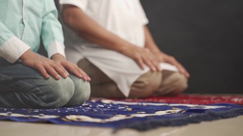 Muslim father and son praying or performing Ssalah while sitting on Prayer rug and touching head to mihrab or mosque printed on rug