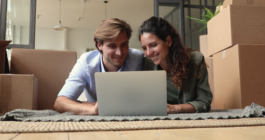 Married couple use laptop choose items decor for new house, search renovation ideas on internet, buy goods for own dwelling feels happy while lying on floor in carpet with underfloor heating system Royalty-Free Stock Footage #1060008482