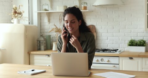 Woman sit at table in kitchen talk on phone to consultant before make order online remotely, businesswoman texting email on laptop chatting with colleague enjoy pleasant conversation working from home