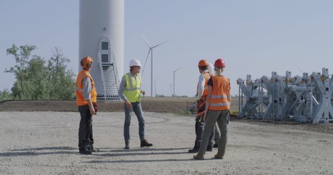 Full length colleagues in vests and helmets examining and discussing construction site of wind farm in countryside