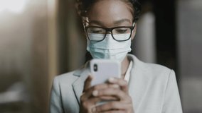 Close-up of young beautiful lady using mobile phone wearing protective mask. Afro-american businesswoman face protection against coronavirus pandemic. Career people. Devices.