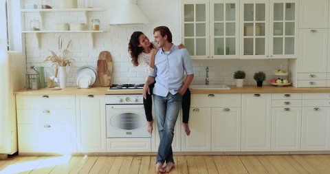 Wide full length view 30s wife sit on kitchen counter top hugs beloved standing husband, young family laughing talking spend weekend time at new modern comfort house. Bank loan, happy marriage concept