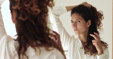 Close up attractive 35s woman in white bath robe standing in bathroom reflected in mirror touch curly brown hair looks pensive thinks about color or hairstyle change, morning routine, haircare concept