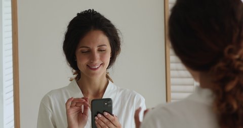 Head shot portrait gorgeous 35s woman in white bath robe standing in cozy bathroom reflected in mirror smiling using smart phone check e-mail. Modern wireless tech easy comfort every day usage concept