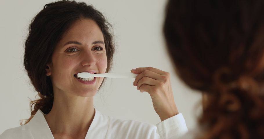 Close up rear back view attractive woman wear bathrobe reflected in mirror holding toothbrush while brush her teeth with toothpaste. Morning routine, dental care, healthcare, personal hygiene concept Royalty-Free Stock Footage #1060020140