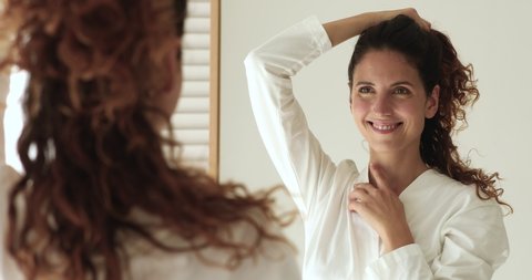 Close up 30s woman in bathrobe freshen up herself in morning looking reflected in mirror touching healthy strong brown curly hairs feels happy. Haircare and styling, best hair products usage concept