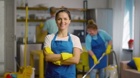 Professional cleaning team in uniform working in modern loft apartment. Portrait of smiling woman janitor with hands crossed posing at camera with colleagues cleaning on background