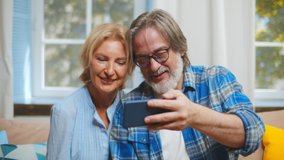 Cheerful mature couple having fun with phone together relaxing on couch together. Portrait of senior husband and wife watching funny video on smartphone and laughing resting at home