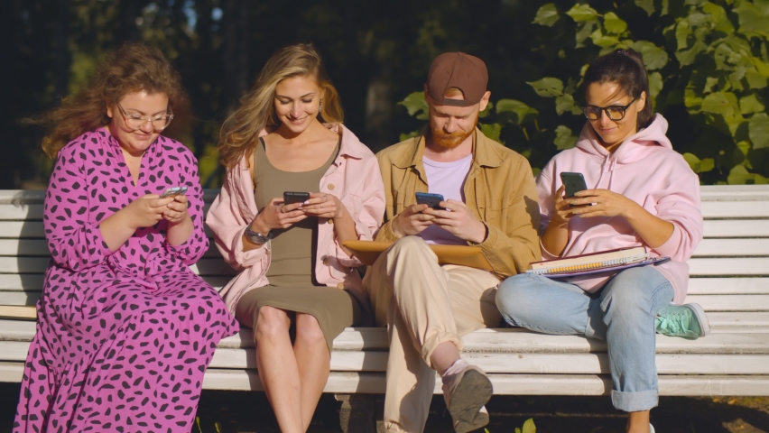 Young man and women sitting on bench outside using mobile app or consulting internet. Four millennial friends surfing internet on smartphone relaxing on bench in park