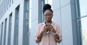 African American happy young businesswoman tapping and scrolling on smartphone. Woman texting message on mobile phone outdoors. Female using gadget and smiling while chatting.
