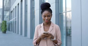 Portrait of African American happy young businesswoman tapping and scrolling on smartphone and smiling to camera. Woman texting message on mobile phone outside. Female using gadget and chatting.