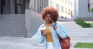 Young stylish African American beautiful curly woman with bag walking at city street, drinking coffee-to-go and smiling joyfully. Pretty happy female strolling outdoors and sipping hot drink.