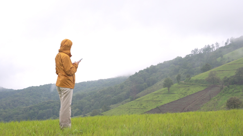 Woman tourist in yellow jacket standing on mountain peak using smartphone with internet for outdoor online working or social media. Female enjoy hiking with wireless technology on travel vacation.