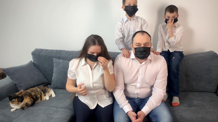 young family, a man, a woman and two sons are sitting in a room on a gray sofa in black viral masks, the concept is a cozy home, love, happy parenthood during a viral epidemic	
