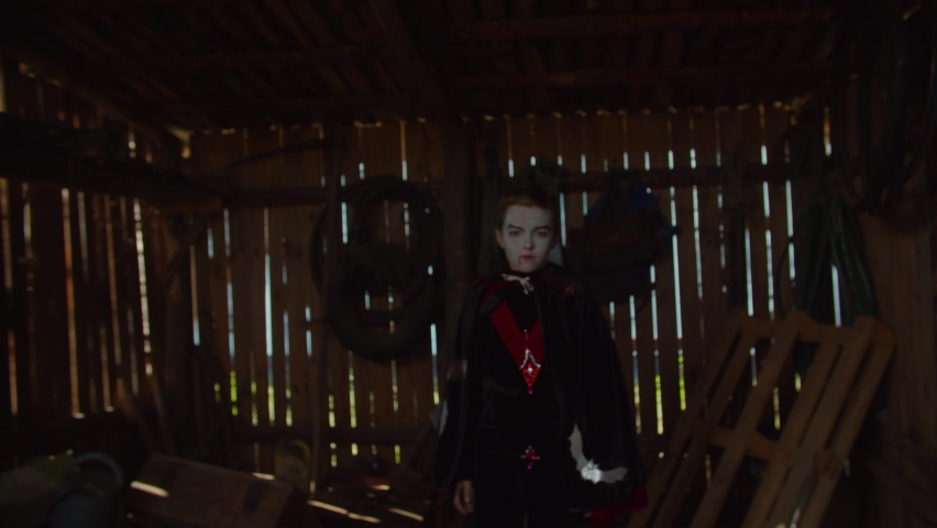 Spooky preadolescent boy with colorful halloween make-up & wearing vampire costume, frightening and furiously whispering at someone in dark rustic shed during halloween holiday.
