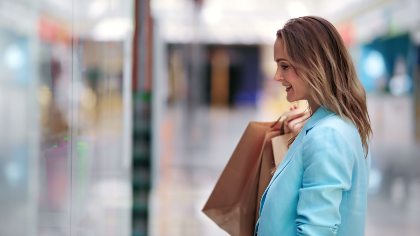 Happy shopaholic female looking at glass showcase at mall center holding paper shopping bags. Charmed stylish woman smiling admiring clothes at seasonal sale. Shot with RED camera in 4K | Shutterstock HD Video #1060038941