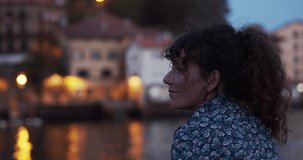 Curly haired woman tourist smiles observing a traditional village in the Basque Country illuminated with lights at night	