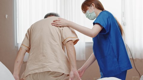 Female Asian nurse support senior male patient stand up and walk from bed in hospital. Nursing home, medical service, physiotherapy, hospitality, or recovery treatment concept