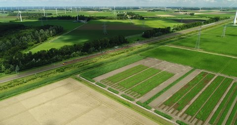 4k aerial horizon view of intercity train passing on railway surrounded by green landscape farmland and electricity pylons with energy generating wind turbines, Flevoland in the Netherlands
