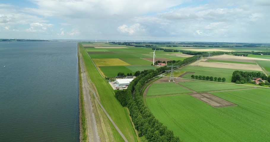 Aerial wide angle view of typical Dutch agricultural landscape along dike with green fields in the foreground blue sky with clouds casting shadows on the land. Wind turbines in the background, Holland Royalty-Free Stock Footage #1060048622