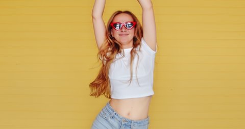 Beautiful hipster unusual woman dancing funny dance on a yellow background. Modern fashionable female in sunglasses fooling around, laughing and grimacing. Teen dancing.