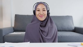Asia muslim lady wear headphone using computer laptop talk to colleagues about sale report in video call while remotely work from home at living room. Social distancing, quarantine for corona virus.