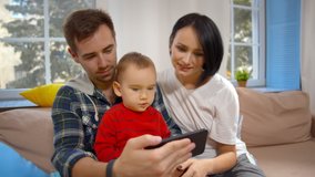 Happy young family with little son using cellphone together. Cheerful mother father and toddler boy sitting on couch and watching cartoon or taking selfie on mobile phone in living room
