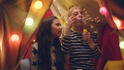 Cute boy and girl having fun in tent with lights blowing soap bubbles. Portrait of caucasian brother and sister wearing pajama at night party in wigwam playing together