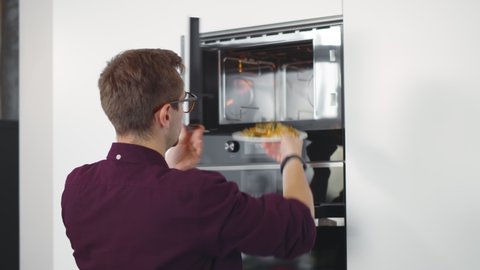 Young man warming up dinner in microwave in home kitchen. Back view of guy putting plate with meal in modern microwave preparing lunch standing in modern kitchen
