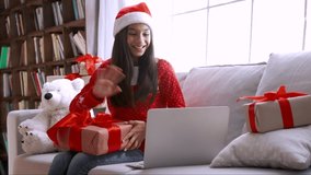 Excited happy indian child kid girl wears santa hat holds xmas gift box, opens Christmas present on computer video call virtual meeting chat with family, social distance friend sits on sofa at home.
