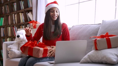 Excited happy indian child kid girl wears santa hat holds xmas gift box, opens Christmas present on computer video call virtual meeting chat with family, social distance friend sits on sofa at home.