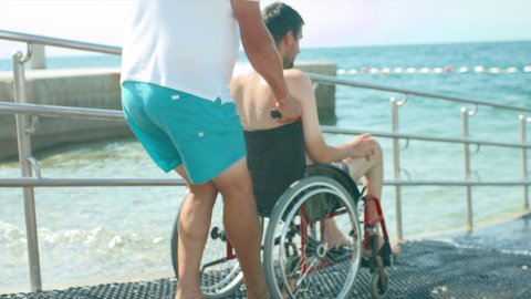 Disabled man at beach going for a swim with help of a wheelchair and assistant.