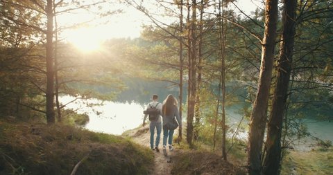 Male female travelers walking hilly trail around mountain lake. Happy couple traveling, go to lakeshore in evening sun light. Follow me, travel together, summer adventure journey in nature outdoors