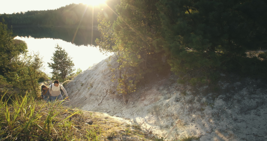 Young male female hikers walking up hilly trail near mountain lake. Couple traveling, hikers go through forest in evening sun light. Travel together, summer adventure journey in nature outdoors
