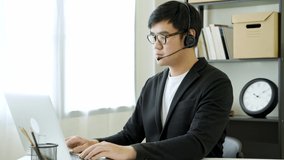 Work from home concept. Asian male business wear a black suit and headphone work at home by video conference explain customer and show sheet sit at home office desk with laptop. 4K Resolution.