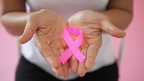 Close up of Hand holding pink ribbon to camera on pink background. World breast cancer day concept. Breast cancer symbol.