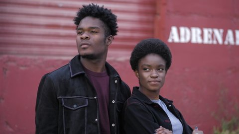 positive African-American couple in stylish black jackets stands near old red garage building on sunny day closeup slow motion