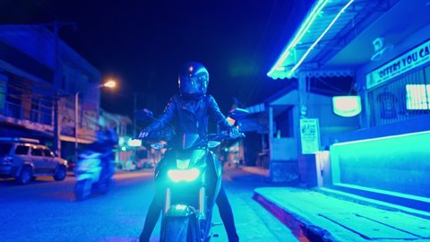Woman wearing a protective helmet, a black leather jacket and pants, sitting on a black motorcycle in blue neon light at night. 4K