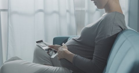 Pregnant mother watching a sonogram of her baby and caressing her belly, she is sitting on the sofa at home