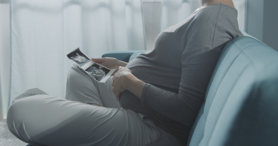 Pregnant woman sitting on the sofa at home and watching ultrasound images of her baby