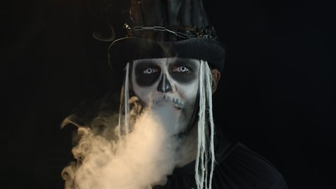 Frightening man in skeleton Halloween cosplay costume. Guy in creepy skull makeup exhaling cigarette smoke from his mouth and smiling. Smoking kills. Day of The Dead. Black background. 6k downscale