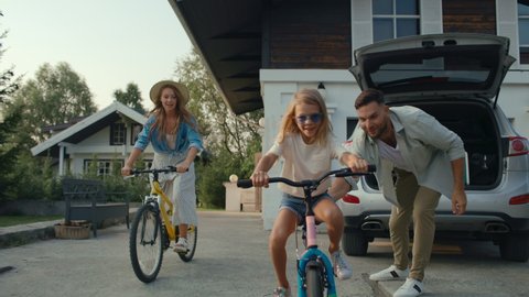 Happy family spends time together on vacation. Attractive mom and pretty daughter having fun outside and riding bikes. Smiling dad in cottage garden got bicycle out of car for leisure of excited girl