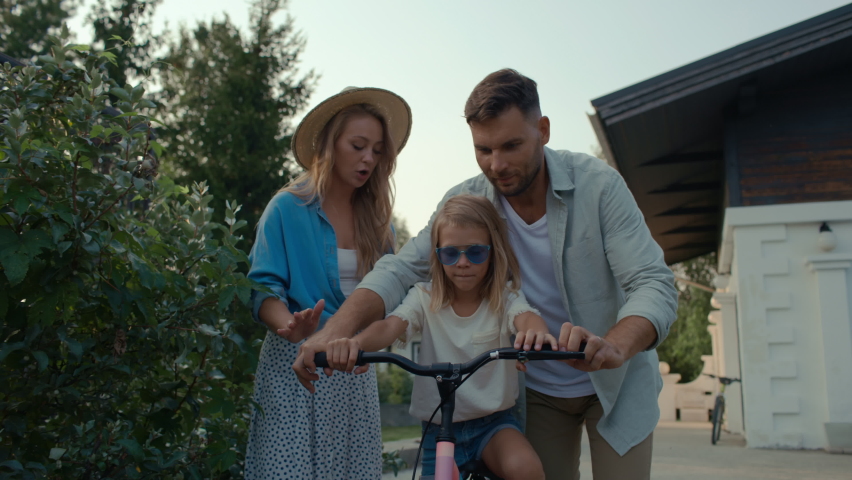 Happy family on vacation spending carefree time together outside. Attractive dad and pretty mom with daughter having fun and walking in nature. Leisure of excited girl riding bicycle in cottage garden Royalty-Free Stock Footage #1060085825