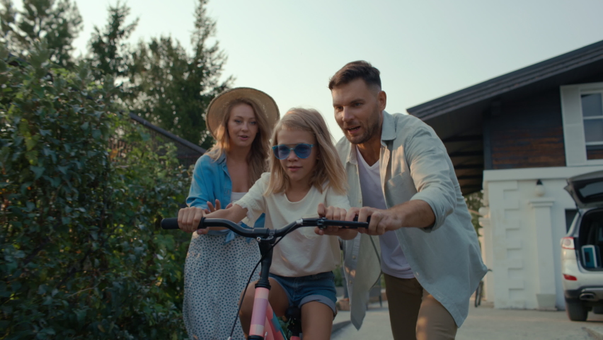 Happy family on vacation spending carefree time together outside. Attractive dad and pretty mom with daughter having fun and walking in nature. Leisure of excited girl riding bicycle in cottage garden | Shutterstock HD Video #1060085825